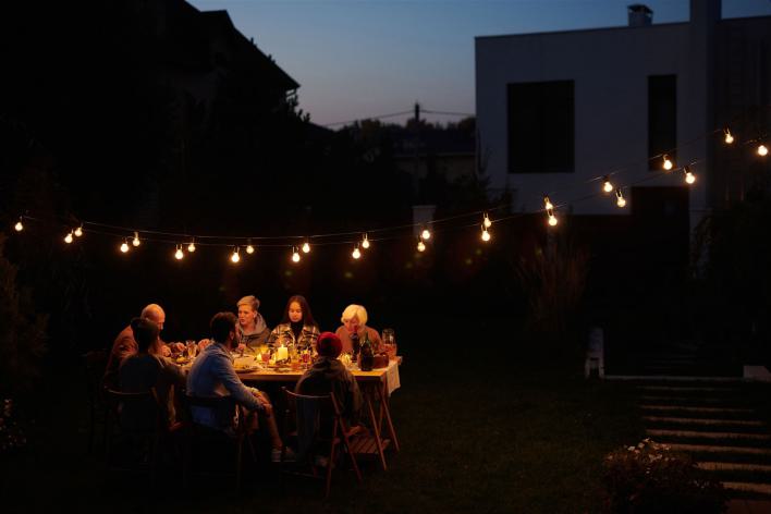 Eine Familie genießt ein Abendessen im Freien unter dekorativen Lichterketten in einem Garten.