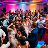 Auf ein KI-generiertes Bild sind festlich gekleidete Menschen, die gerade feiern und tanzen. Buntes Licht, das DJ Equipment & die passende Musik sorgen für die perfekte Stimmung. 